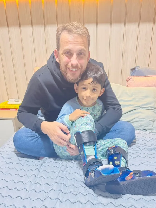 Gabriel sentado no colo do pai, usando a órtese azul nas pernas
