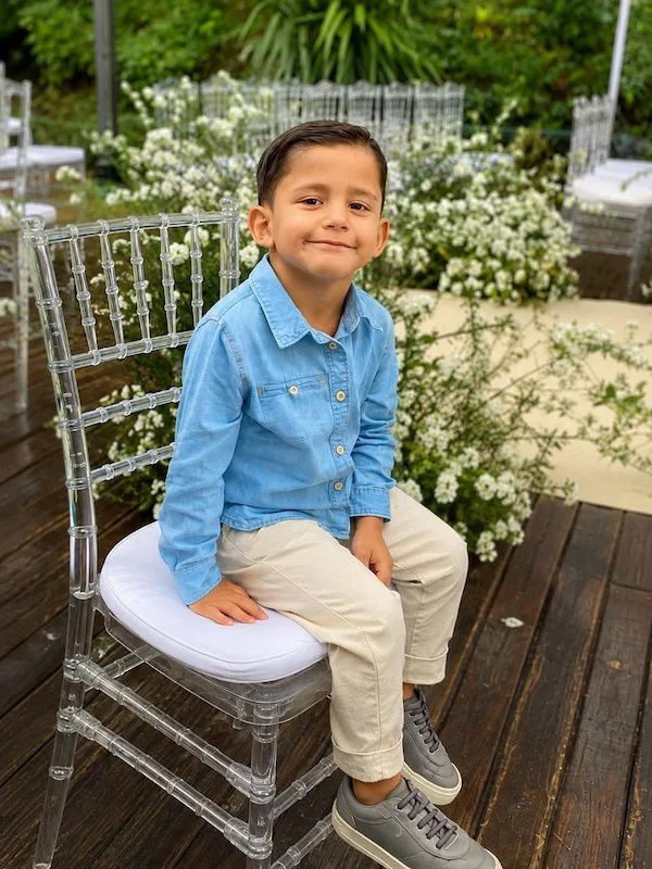 Gabriel sorrindo, sentado numa cadeira, de camisa jeans azul e calça bege